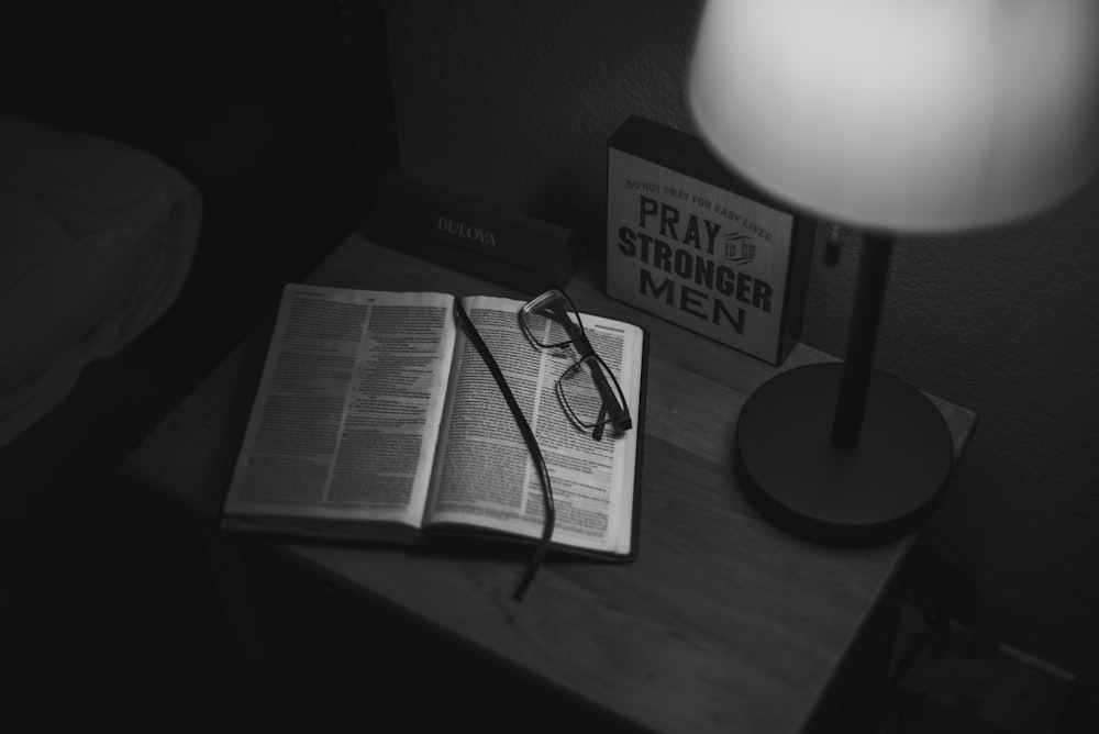 grayscale photography of eyeglasses on open book near table lamp