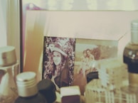 A framed photograph rests on a surface surrounded by various bottles and containers. The first image in the frame depicts a person in a hat with a backdrop of foliage. The second photo shows two individuals, possibly a couple, sitting together with a scenic outdoor setting in the background. The overall setting appears to be nostalgic and personal.