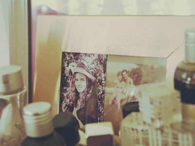 A framed photograph rests on a surface surrounded by various bottles and containers. The first image in the frame depicts a person in a hat with a backdrop of foliage. The second photo shows two individuals, possibly a couple, sitting together with a scenic outdoor setting in the background. The overall setting appears to be nostalgic and personal.