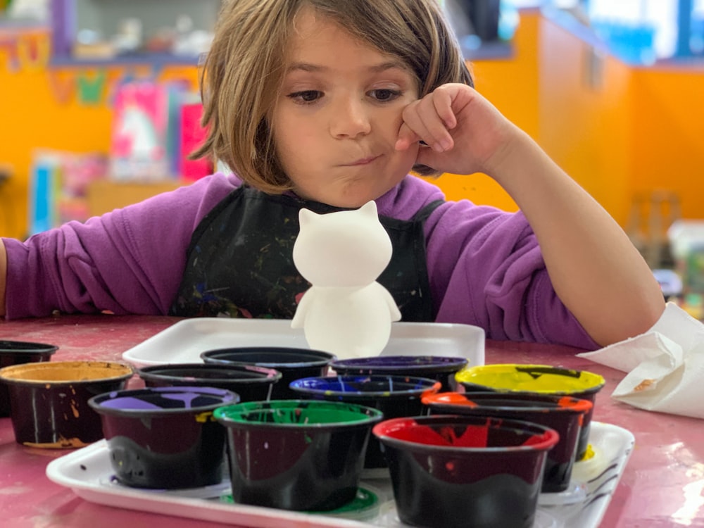 criança sentada na frente da mesa com brinquedo animal branco e recipientes de tintas