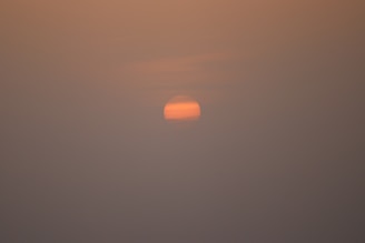 A dim, pinkish-orange sun is partially obscured by layers of haze or clouds, creating a muted and serene atmosphere. The surrounding sky is filled with soft gradients of orange and gray, suggesting low visibility and a calm setting.