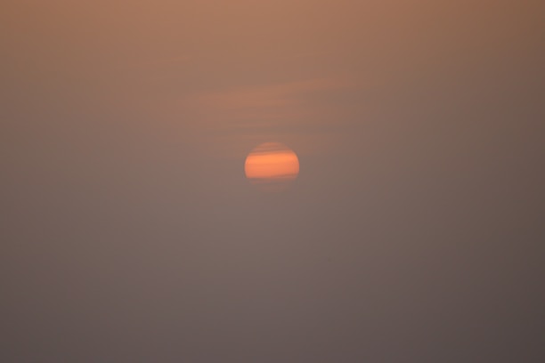 A dim, pinkish-orange sun is partially obscured by layers of haze or clouds, creating a muted and serene atmosphere. The surrounding sky is filled with soft gradients of orange and gray, suggesting low visibility and a calm setting.