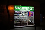 A brightly lit convenience store with a prominent green sign reading 'Supermercat Botiga de Conveniencia.' The store window displays an array of merchandise, including plates, souvenirs, and items featuring the FC Barcelona logo.