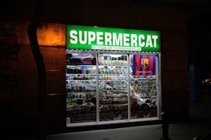 A brightly lit convenience store with a prominent green sign reading 'Supermercat Botiga de Conveniencia.' The store window displays an array of merchandise, including plates, souvenirs, and items featuring the FC Barcelona logo.