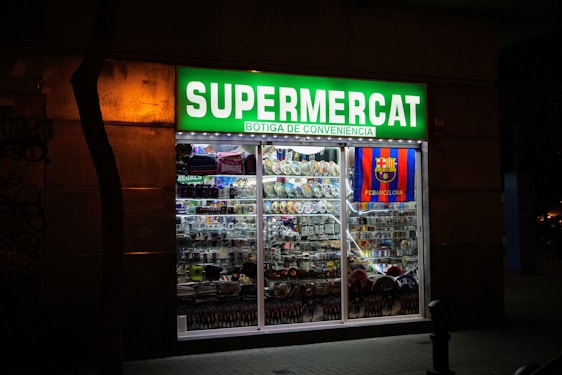 A brightly lit convenience store with a prominent green sign reading 'Supermercat Botiga de Conveniencia.' The store window displays an array of merchandise, including plates, souvenirs, and items featuring the FC Barcelona logo.