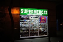 A brightly lit convenience store with a prominent green sign reading 'Supermercat Botiga de Conveniencia.' The store window displays an array of merchandise, including plates, souvenirs, and items featuring the FC Barcelona logo.