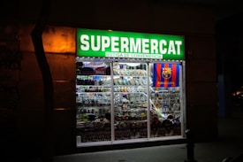 A brightly lit convenience store with a prominent green sign reading 'Supermercat Botiga de Conveniencia.' The store window displays an array of merchandise, including plates, souvenirs, and items featuring the FC Barcelona logo.