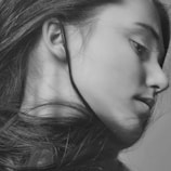 A grayscale portrait of a person focusing on the side profile, with long flowing hair. The portrait is detailed, capturing subtle features such as the texture of the skin and individual strands of hair. The lighting creates gentle shadows and highlights, emphasizing the contours of the face.