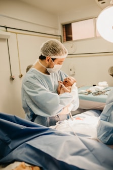 A healthcare professional wearing a mask and hairnet holds a newborn baby in a medical setting. The baby is crying while the person looks down at them. The environment appears to be a hospital or a surgical room, with medical equipment and another person partially visible in the background.