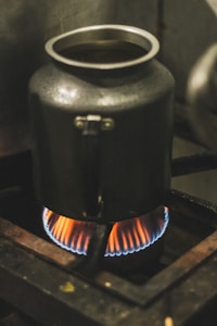 A metal container is placed on a gas stove with a visible blue and orange flame underneath, indicating it is being heated. The surrounding area suggests an industrial or heavily used kitchen setting.