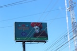 A large billboard advertising a 'Festival de Día de Muertos' features an image of a person with a traditional Day of the Dead face paint and wearing vibrant red marigold flowers in their hair. The background is teal with decorative patterns and Spanish text. Visible power lines and a clear blue sky can be seen.