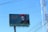 A large billboard advertising a 'Festival de Día de Muertos' features an image of a person with a traditional Day of the Dead face paint and wearing vibrant red marigold flowers in their hair. The background is teal with decorative patterns and Spanish text. Visible power lines and a clear blue sky can be seen.