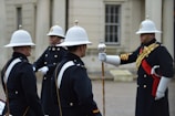 A group of uniformed soldiers, wearing white helmets and dark formal attire with gold and red details, stand in formation. One of the soldiers holds a ceremonial staff and seems to be giving instructions. The background features a historic building with white stone walls and traditional windows.