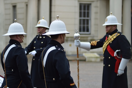 A group of uniformed soldiers, wearing white helmets and dark formal attire with gold and red details, stand in formation. One of the soldiers holds a ceremonial staff and seems to be giving instructions. The background features a historic building with white stone walls and traditional windows.