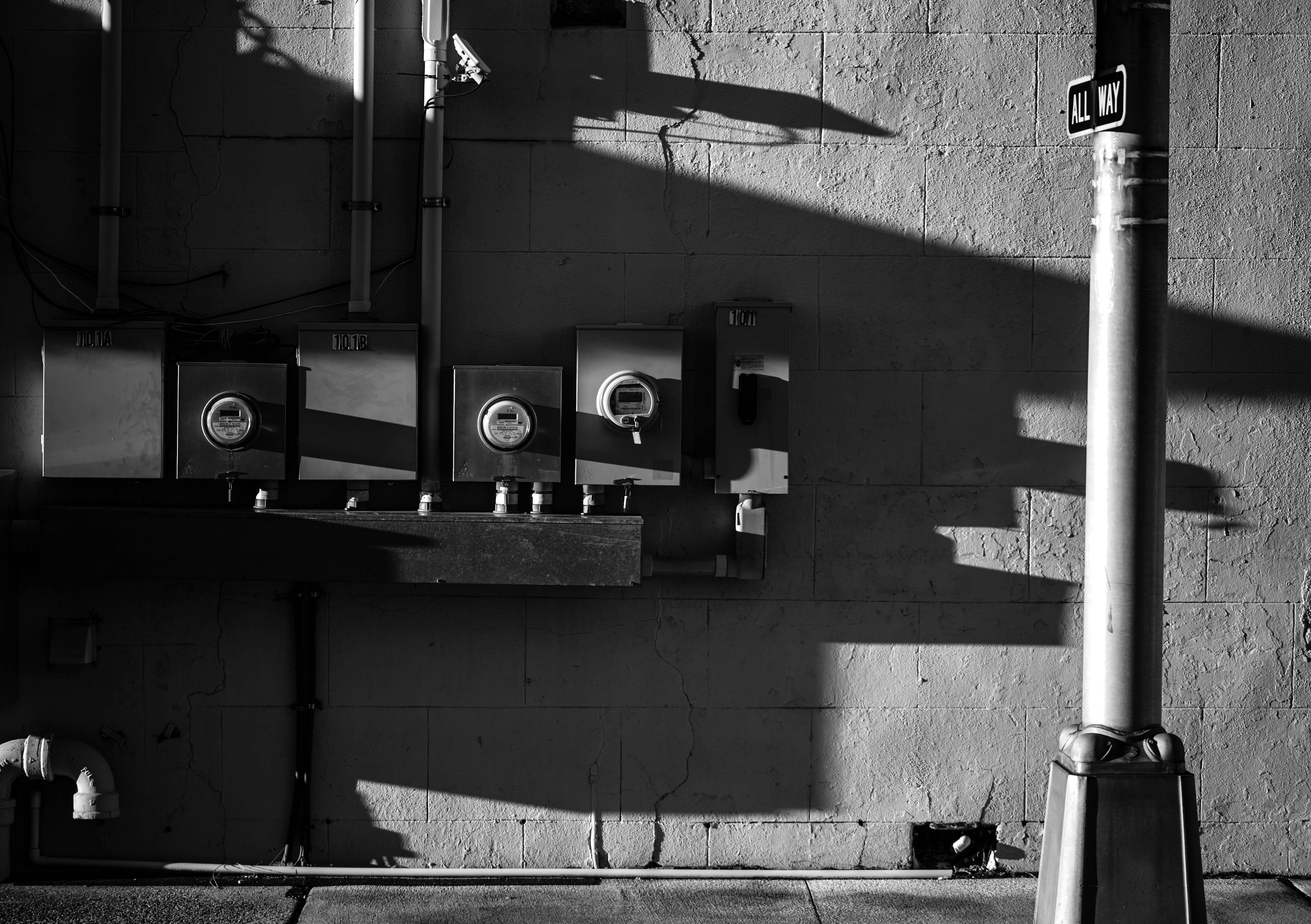 A black and white image depicting a wall with three electrical meters and a few pipes extending vertically. Shadows from a nearby structure create angles across the wall's surface, adding texture. On the right, a large metal pole extends vertically with an 'All Way' street sign attached near the top. The wall appears worn, with visible cracks and rough textures.