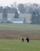 A rural landscape featuring a field with three people dressed in dark clothing walking across it. In the background, there is a silo and a barn surrounded by trees, with a slightly overcast sky.