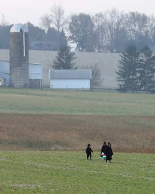 A rural landscape featuring a field with three people dressed in dark clothing walking across it. In the background, there is a silo and a barn surrounded by trees, with a slightly overcast sky.