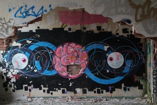 A vibrant and detailed graffiti artwork on a dilapidated wall. The central focus is a large pink brain surrounded by blue concentric circles, flanked by two large white eyes with pink pupils. The wall is partially destroyed, with chipped paint and exposed bricks, emphasizing an urban decay environment.
