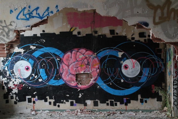 A vibrant and detailed graffiti artwork on a dilapidated wall. The central focus is a large pink brain surrounded by blue concentric circles, flanked by two large white eyes with pink pupils. The wall is partially destroyed, with chipped paint and exposed bricks, emphasizing an urban decay environment.