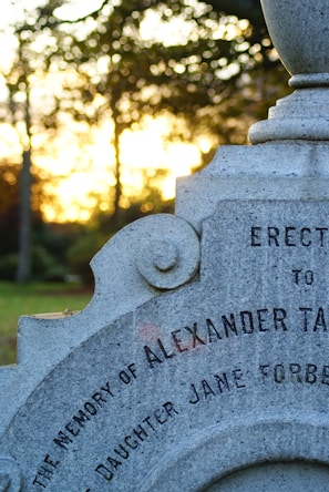 A close-up of an old, weathered tombstone with engraved text commemorating Alexander and his daughter Jane Forbes. The background features blurred trees and a golden sunset sky, creating a serene and contemplative atmosphere.