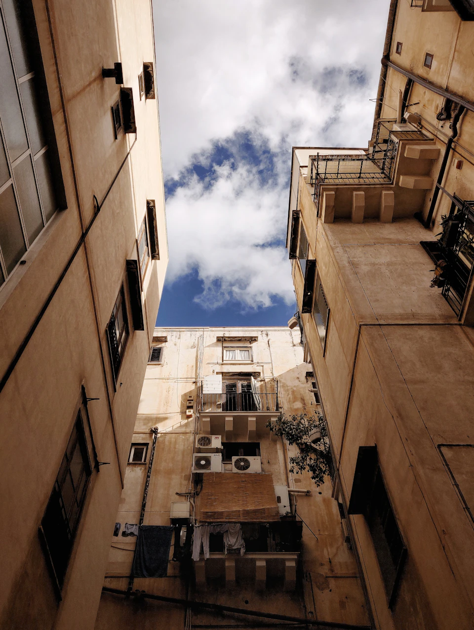 Two buildings flank a narrow street, one beige and one brown, with balconies and trees