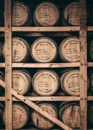 Wooden barrels stacked on a wooden rack, each labeled with 'Kentucky Peerless Distilling Co', indicating the contents as rye, along with serial and batch numbers. The barrels are arranged closely within a rustic storage setting.