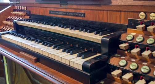 A vintage pipe organ keyboard featuring multiple rows of keys and numerous stops and switches on either side. The wood finish exudes a classic and elegant look, with polished surfaces and intricately designed components.