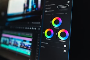 A close-up view of a digital video editing software interface, focusing on color grading tools. Three color wheels labeled Midtones, Shadows, and Highlights are prominent, with sliders for adjusting color balance. The background shows a blurred timeline with video and audio tracks, indicating an ongoing editing process.