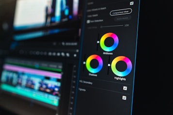 A close-up view of a digital video editing software interface, focusing on color grading tools. Three color wheels labeled Midtones, Shadows, and Highlights are prominent, with sliders for adjusting color balance. The background shows a blurred timeline with video and audio tracks, indicating an ongoing editing process.