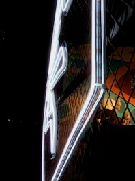 A brightly illuminated neon sign viewed from the side against a dark background. The sign appears to be enclosed in a mesh, creating a pattern of shadows. Behind the mesh, a collection of plush toys, including a teddy bear, adds a playful touch to the scene.