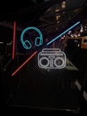 Neon outlines of headphones and a boombox are set against a dark, mesh-like background. The neon colors are bright, with vivid blue, red, and white elements. The setting appears to be an evening or nighttime environment with some ambient lighting in the distance.