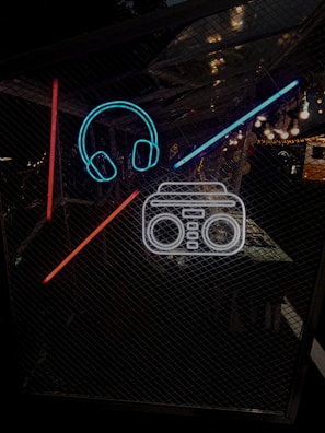 Neon outlines of headphones and a boombox are set against a dark, mesh-like background. The neon colors are bright, with vivid blue, red, and white elements. The setting appears to be an evening or nighttime environment with some ambient lighting in the distance.