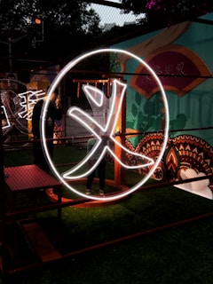 A large illuminated Chinese character is displayed within a circular frame. The scene is set against a vibrant mural featuring various colors and patterns. The character is outlined in bright white neon, giving it a striking appearance in the dimly lit environment. There is greenery at the bottom, and the setting appears to be an artistic or cultural installation.