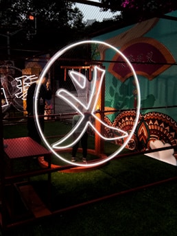 A large illuminated Chinese character is displayed within a circular frame. The scene is set against a vibrant mural featuring various colors and patterns. The character is outlined in bright white neon, giving it a striking appearance in the dimly lit environment. There is greenery at the bottom, and the setting appears to be an artistic or cultural installation.