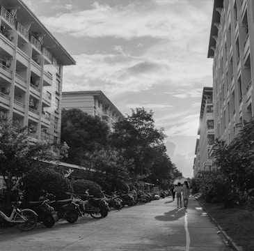 A black and white urban scene showing a walkway lined with bicycles and mopeds. Tall residential buildings frame both sides of the path, with trees and bushes alongside. People are walking down the path, including a pair with an umbrella. The sky is partly cloudy, adding texture to the background.