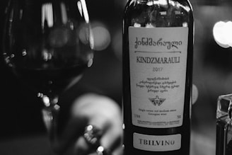 A close-up of a wine bottle labeled 'Kindzmarauli 2017' with a focus on the intricate design and text on the label. Next to the bottle, a glass of wine and part of a person's hand holding the glass can be seen. The image is in black and white, giving it a classic and elegant appearance.