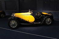 A vintage car with a bright yellow body and black fenders is displayed in a dimly lit room. The vehicle features sleek curves, large silver wheels, and a shiny chrome grille and headlights.