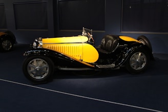 A vintage car with a bright yellow body and black fenders is displayed in a dimly lit room. The vehicle features sleek curves, large silver wheels, and a shiny chrome grille and headlights.