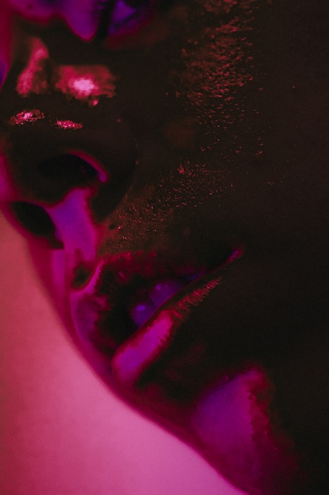 A close-up of a person's face with a focus on the lips and surrounding skin, illuminated by dramatic pink and purple lighting, creating deep shadows and highlights.