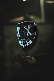 A glowing mask with neon blue outlines forms a face with Xs over the eyes and a grid-like pattern for a mouth. The mask appears to be floating above an outstretched hand, set against a dark and blurred background, adding a mysterious atmosphere.