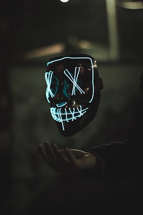 A glowing mask with neon blue outlines forms a face with Xs over the eyes and a grid-like pattern for a mouth. The mask appears to be floating above an outstretched hand, set against a dark and blurred background, adding a mysterious atmosphere.