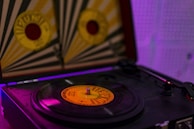 A close-up of a vintage record player with a vinyl record spinning on the turntable. The setup features a distinctive record cover with a yellow and red design in the background, while the room is dimly lit, casting a purple hue. The tonearm is visible resting on the player.