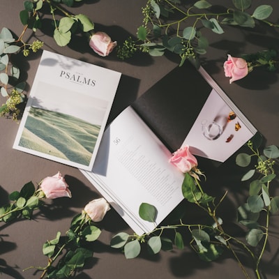 An open book with the title 'Psalms' on the cover lies on a surface surrounded by pink roses and green foliage. The book's pages are open, showing text and an image of a glass vase with flower petals. The composition is artistic and serene, suggesting a theme of peace and reflection.