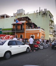 A bustling street scene with a large crowd gathered outside an orange two-story building adorned with Puerto Rican flags. People are socializing around parked cars, motorcycles, and a scooter. The building has a balcony, and some people are observing from above. The setting conveys a lively urban atmosphere.