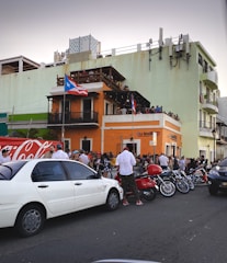 A bustling street scene with a large crowd gathered outside an orange two-story building adorned with Puerto Rican flags. People are socializing around parked cars, motorcycles, and a scooter. The building has a balcony, and some people are observing from above. The setting conveys a lively urban atmosphere.