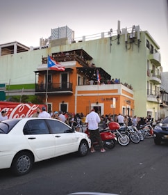 A bustling street scene with a large crowd gathered outside an orange two-story building adorned with Puerto Rican flags. People are socializing around parked cars, motorcycles, and a scooter. The building has a balcony, and some people are observing from above. The setting conveys a lively urban atmosphere.