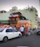 A bustling street scene with a large crowd gathered outside an orange two-story building adorned with Puerto Rican flags. People are socializing around parked cars, motorcycles, and a scooter. The building has a balcony, and some people are observing from above. The setting conveys a lively urban atmosphere.