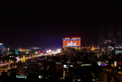 A vibrant cityscape at night with a prominent building displaying a large, colorful digital screen. The skyline features illuminated structures, and the streets below are bustling with bright lights and traffic.