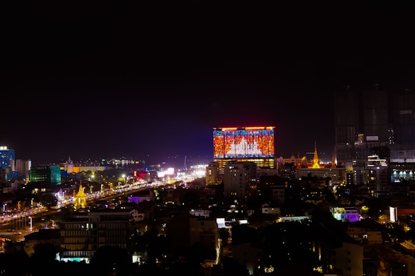 A vibrant cityscape at night with a prominent building displaying a large, colorful digital screen. The skyline features illuminated structures, and the streets below are bustling with bright lights and traffic.