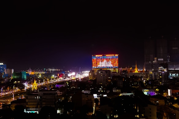 A vibrant cityscape at night with a prominent building displaying a large, colorful digital screen. The skyline features illuminated structures, and the streets below are bustling with bright lights and traffic.
